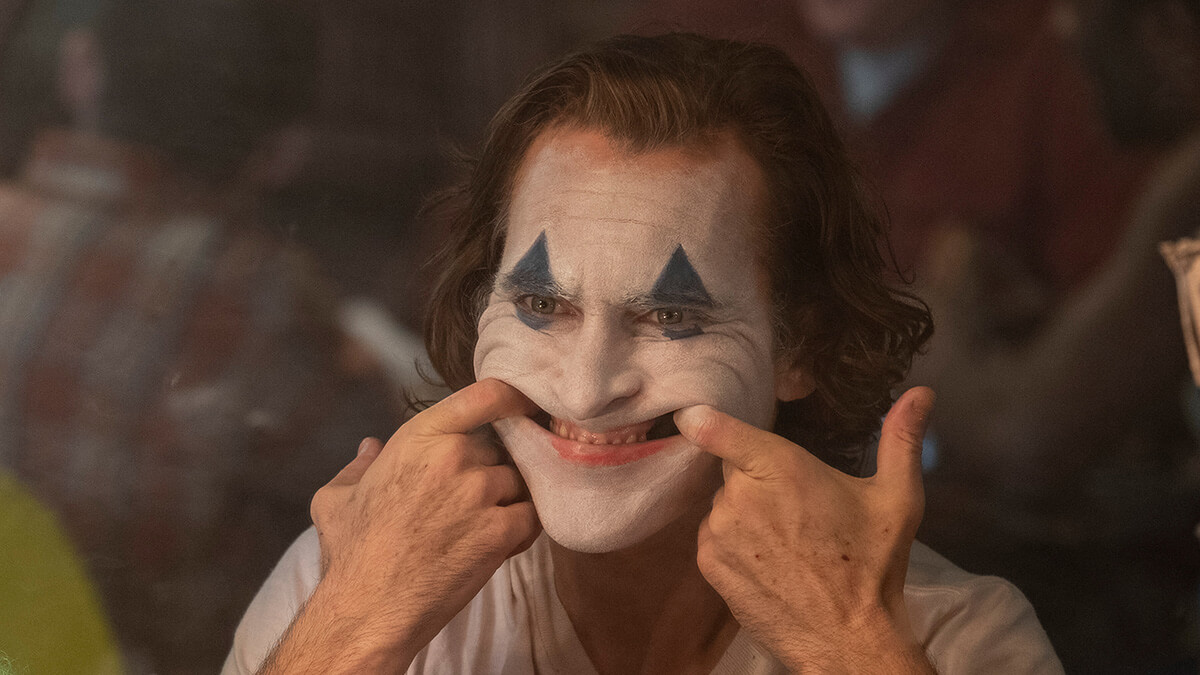 Joker meletakkan jarinya ke mulut untuk membuat senyum di wajahnya.
